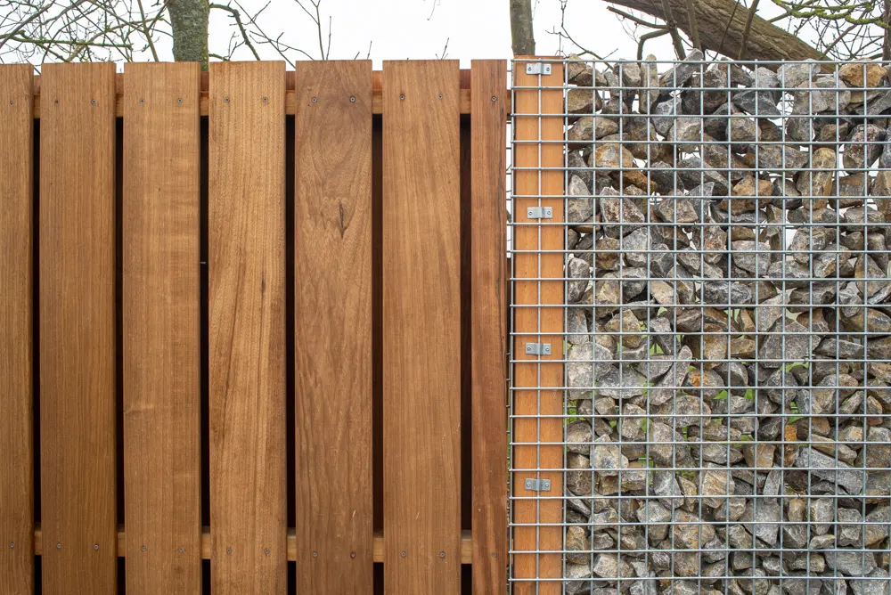 Installed Yellow Balau timber post supporting a vertical wooden slat privacy fence panel situated adjacent to a wire mesh gabion stone wall.