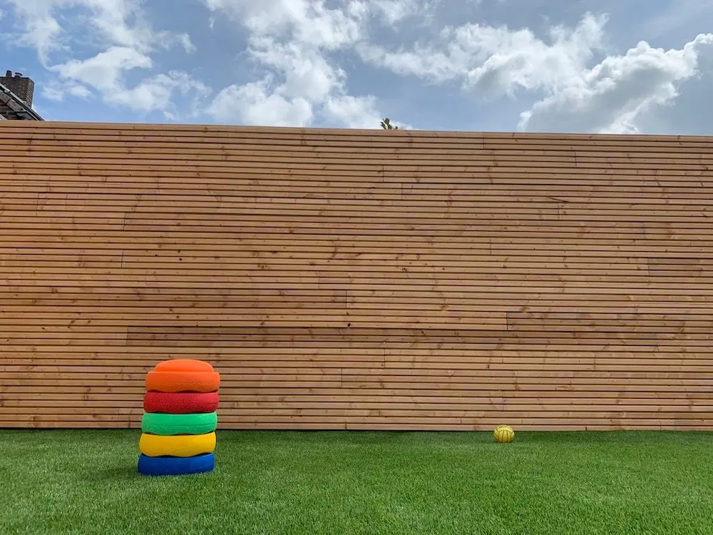 Installed Ecowood triple slatted profile cladding boards creating a large, seamless natural wood boundary wall behind an artificial lawn.