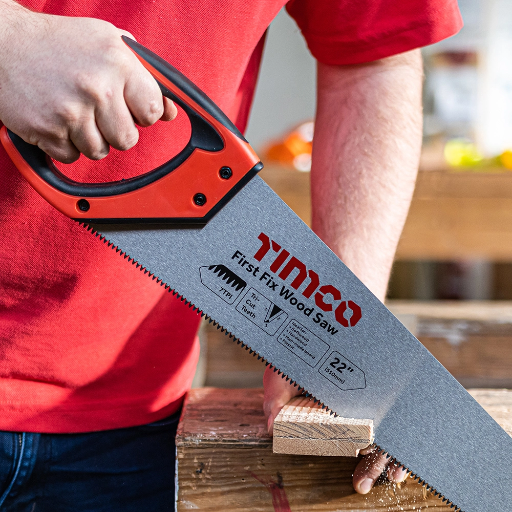 A close-up action view of a worker using a TIMCO second Fix 22-inch wood saw with a red and black handle to cut through a piece of timber timber on a workbench.